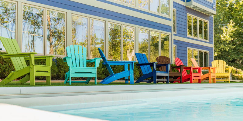A vibrant row of POLYWOOD Adirondack chairs in green, turquoise, blue, navy, brown, red, orange, and yellow, arranged on a patio in front of a modern blue-sided home with large windows, showcasing the brand's colorful and durable outdoor furniture.