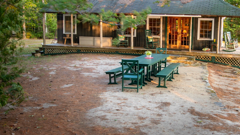 An assortment of green POLYWOOD dining furniture sits in front of a lakeside cabin surrounded by evergreen trees.