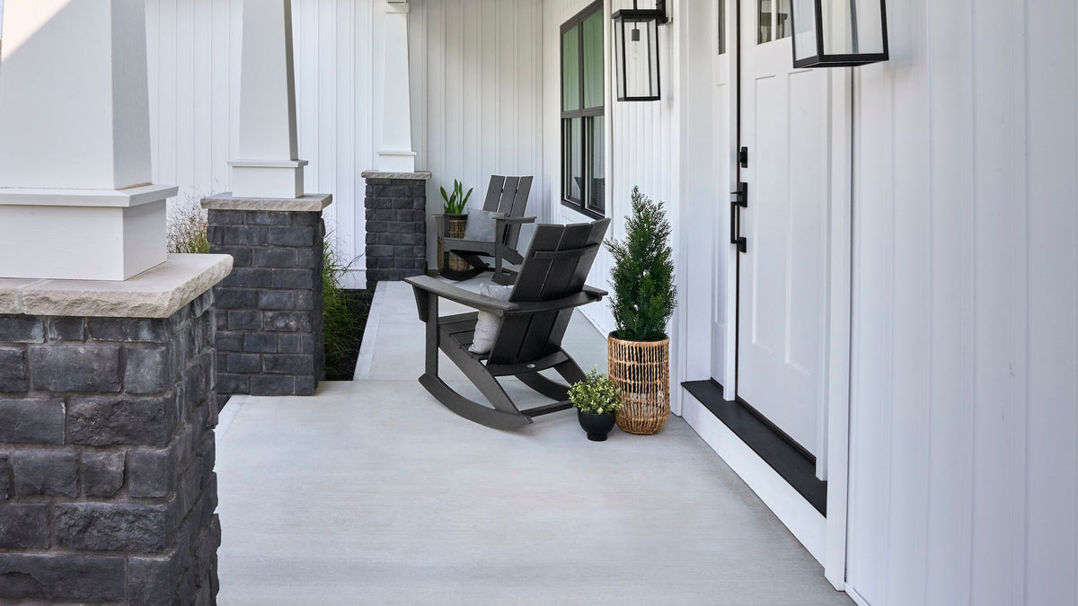 Two POLYWOOD Modern Adirondack Rockers in Black sit on a concrete porch before a white house with grey brick pillars.