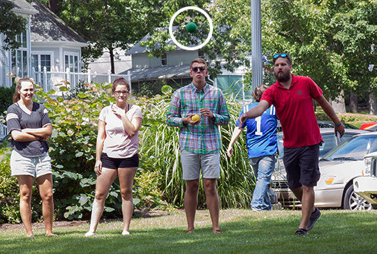 Four people stand outside in front of bushes, one tosses a bocce ball.