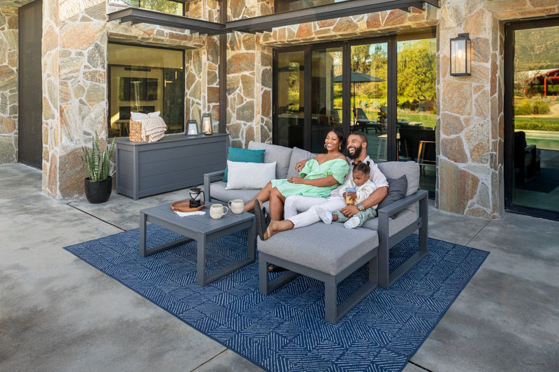 A family sits together on a grey POLYWOOD EDGE 4 Piece Modular Deep Seating Set with Ottoman on a patio behind a stone house.