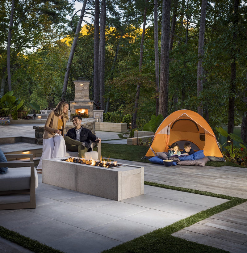 A couple sits by a modern fire pit while two children read in an orange tent on a spacious, wooded patio.