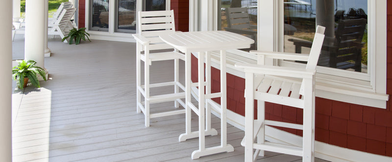 A white POLYWOOD Captain Bar Chair Duo and a Nautical 31 Inch Bar Table sit on a rounded porch of a red house.