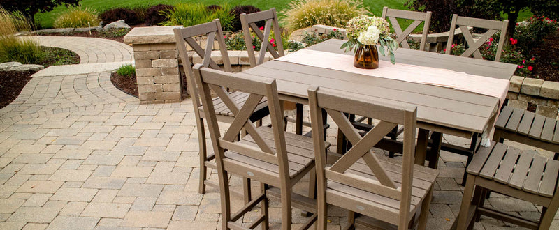 A brown POLYWOOD Braxton 9 Piece Farmhouse Trestle Bar Set with a beige runner and vase of flowers sits on a brick patio.
