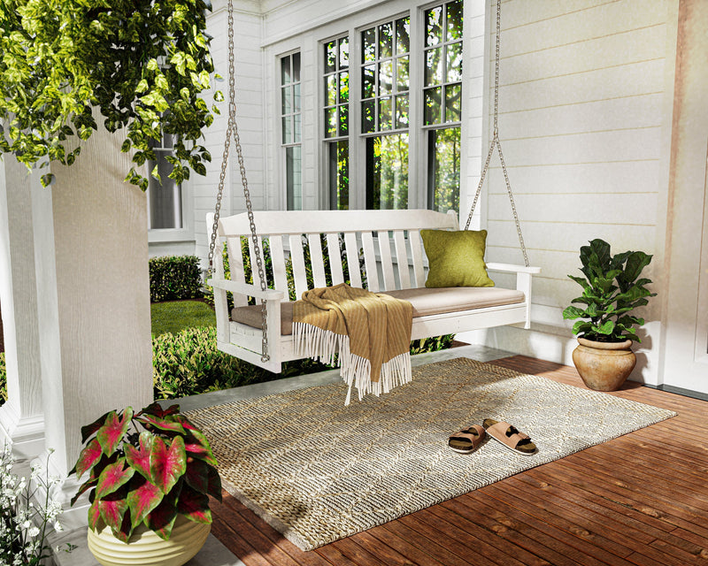 A white POLYWOOD Cottage 60 Inch Swing hangs over a patterned rug on a white house’s wooden porch with potted plants nearby.