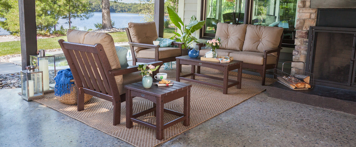 A brown POLYWOOD Vineyard 5-Piece Deep Seating Set with cream cushions on a patio overlooking a lake. Lanterns and a fireplace add to the cozy atmosphere.