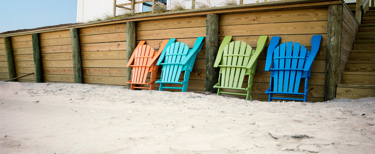 Colorful POLYWOOD Palm Coast Folding Adirondack chairs lean against a wooden wall on a sandy beach.