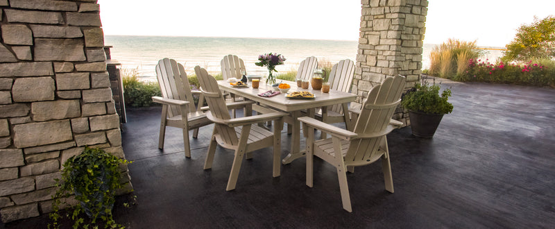 A Vineyard Curveback Adirondack 7 Piece Dining Set with Trestle Legs in Sand sits on a covered oceanside patio.
