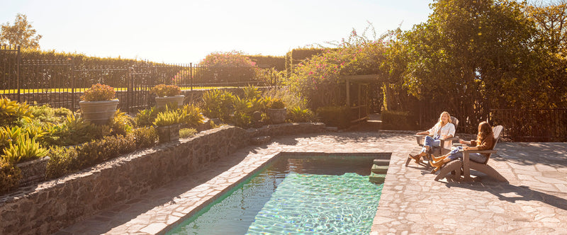 People sit by a swimming pool on tan POLYWOOD Adirondack chairs and ottomans.