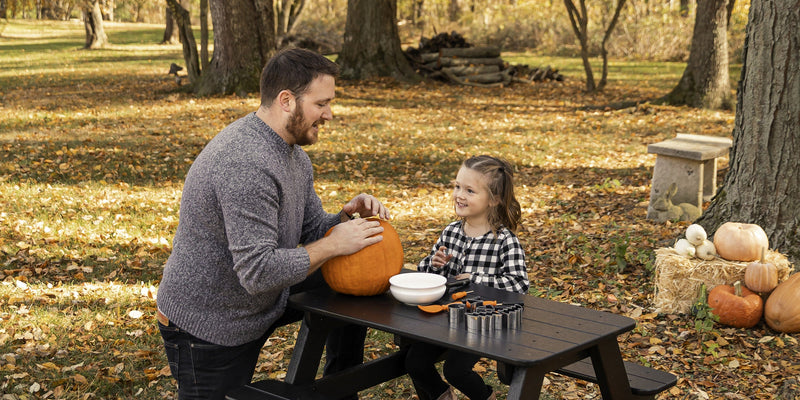 A father and daughter carve a pumpkin together at a black POLYWOOD Kids Picnic Table on a lawn covered in leaves.