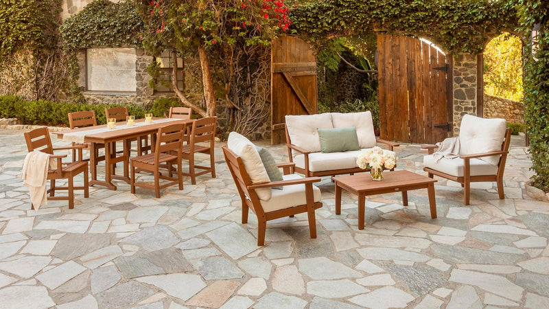 POLYWOOD Lakeside dining and deep seating sets in Teak and Natural Linen sit on a stone patio before a house covered in ivy.