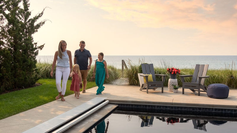 A family of four walks along a poolside patio at sunset, with Adirondack chairs and a table set up near tall grass and a scenic ocean view in the background.