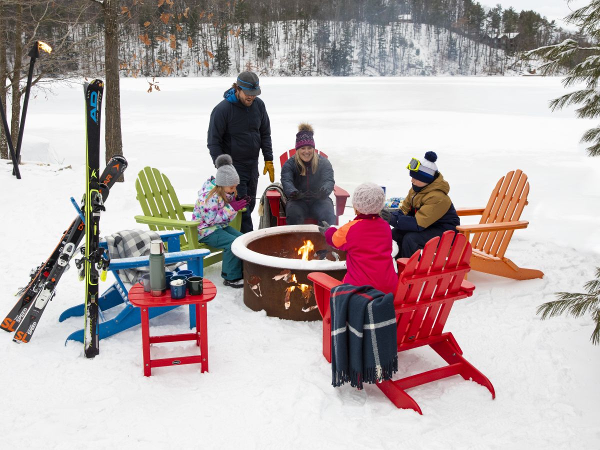 A family sits on colorful POLYWOOD Adirondack chairs around a fire pit near trees and on a snowy day.