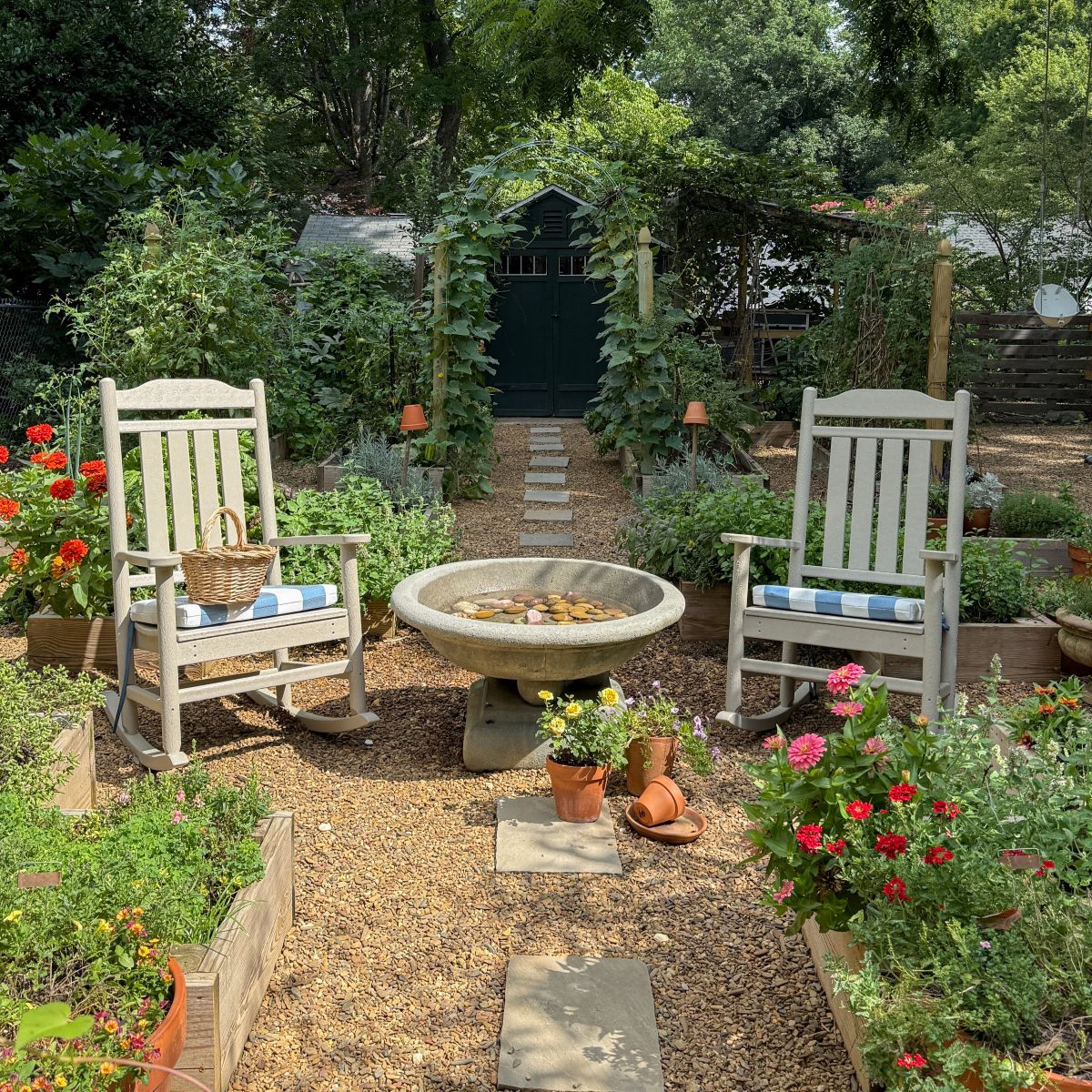 Tan POLYWOOD Cottage Legacy Rocking Chairs sit on a pea gravel path in a raised bed garden filled with blooming flowers..