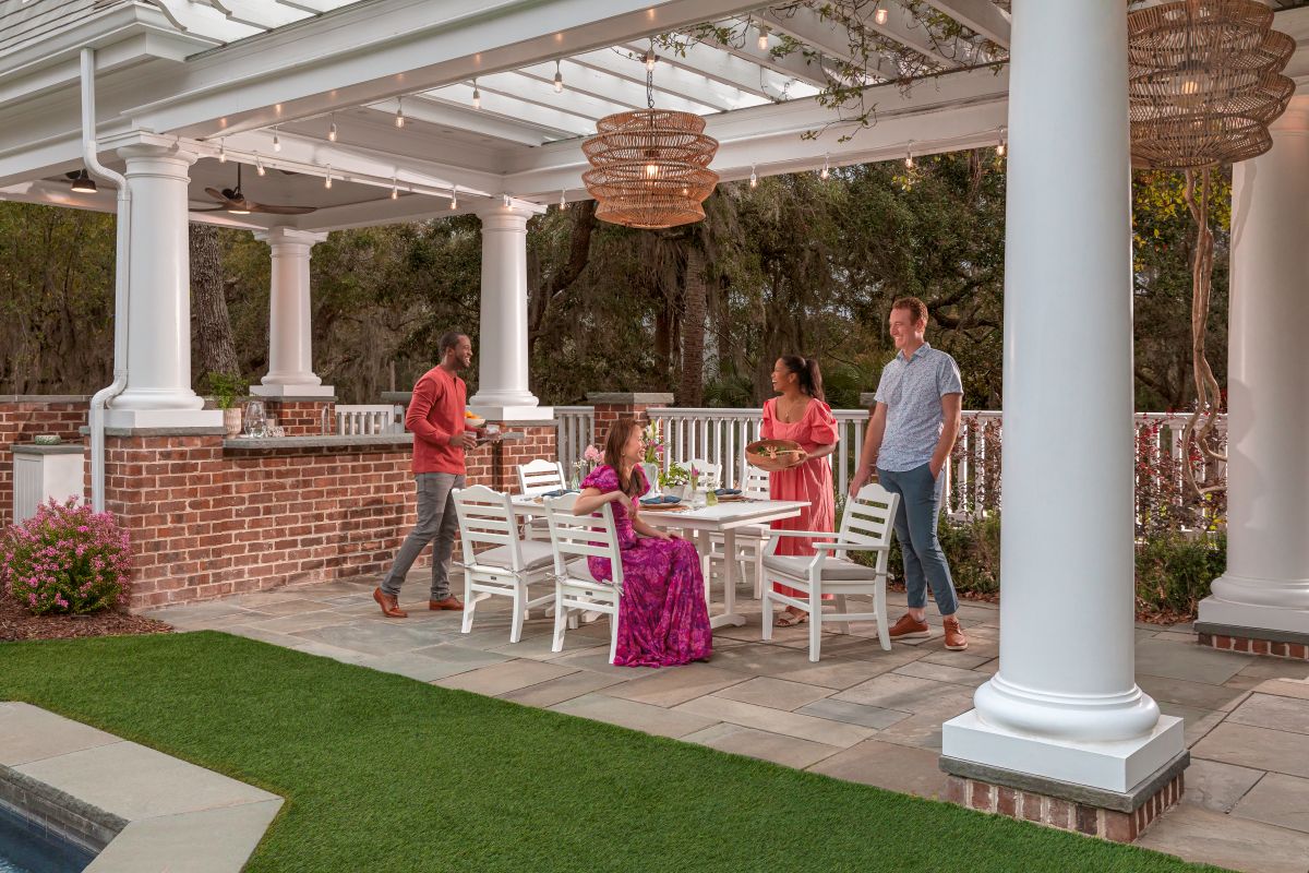 Four adults gather and chat around a white POLYWOOD Savannah 7 Piece Farmhouse Dining Set on a patio under a pergola.