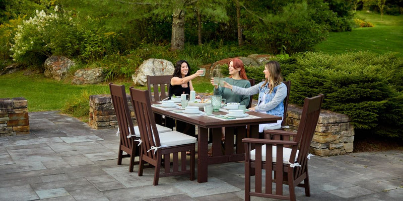 Three women seated around a dark wood outdoor dining table, enjoying drinks and conversation on a stone patio surrounded by greenery.