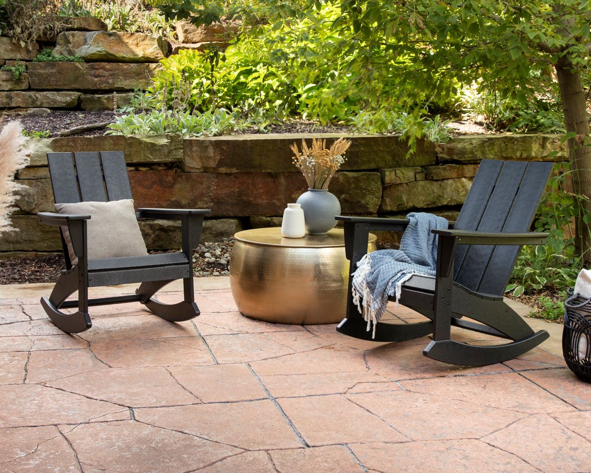 Black POLYWOOD Modern Adirondack Rocking Chairs flank a metallic round table before a stone wall on a flagstone patio.