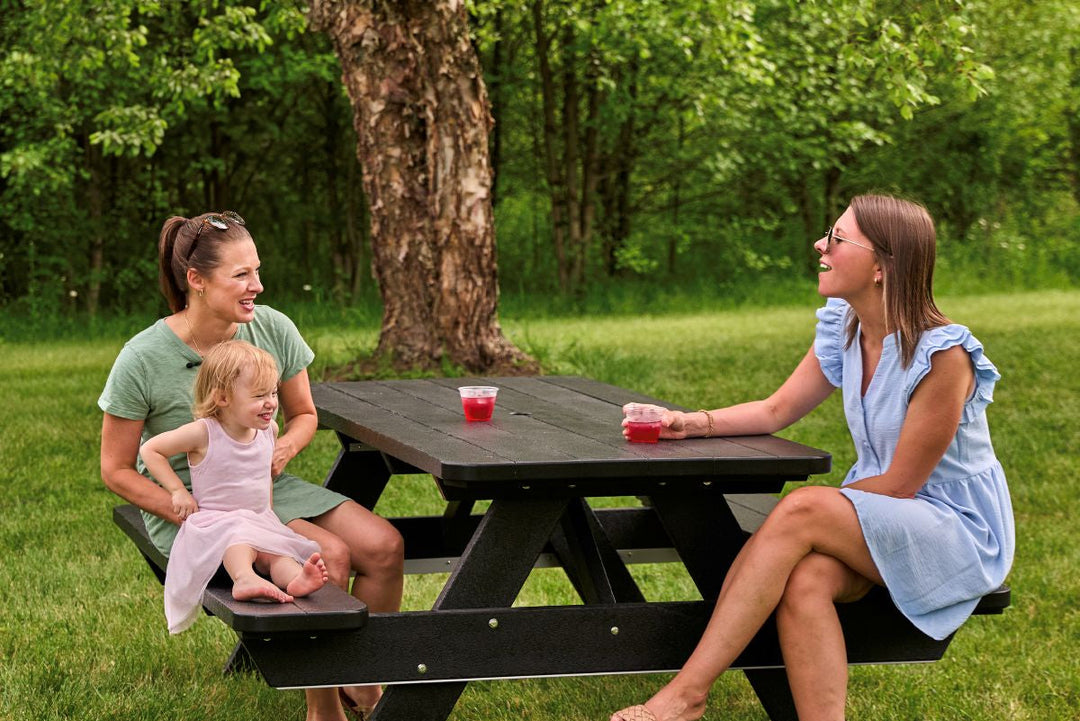 Two women and a child sit at a black POLYWOOD Park 72 Inch Picnic Table on a grassy lawn by trees, laughing over drinks.