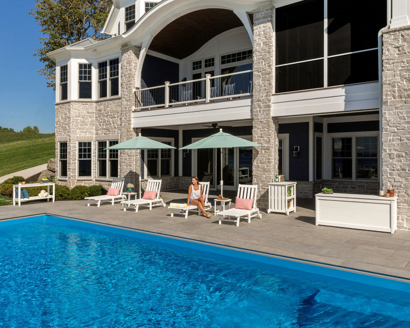 Assorted white POLYWOOD chaise lounges, tables, and umbrellas are situated on a poolside patio before a stone house.