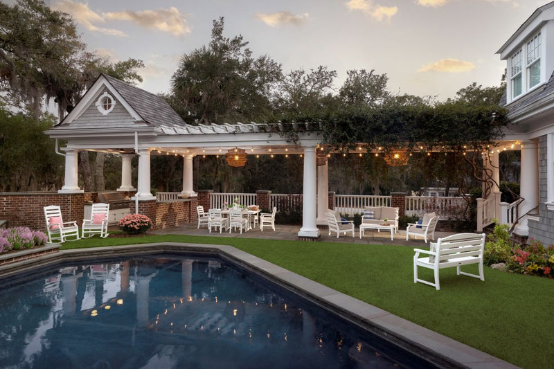 Assorted white POLYWOOD Savannah lounge, dining, and deep seating furniture sits under a pergola with lights by a pool.