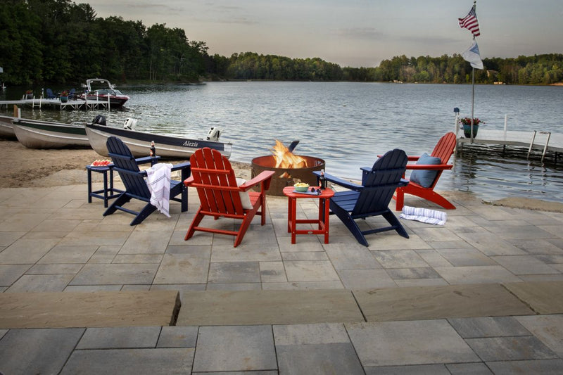 Red and blue POLYWOOD Vineyard Adirondack Chairs sit around a fire pit on a large patio next to a lake surrounded by trees.