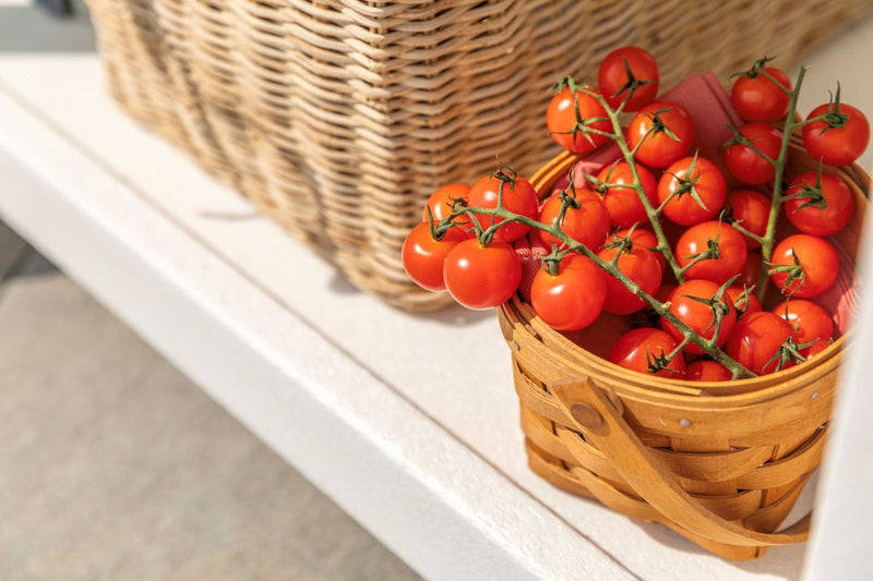 A small wooden basket filled with ripe cherry tomatoes sits on a white POLYWOOD table beside a wicker basket.