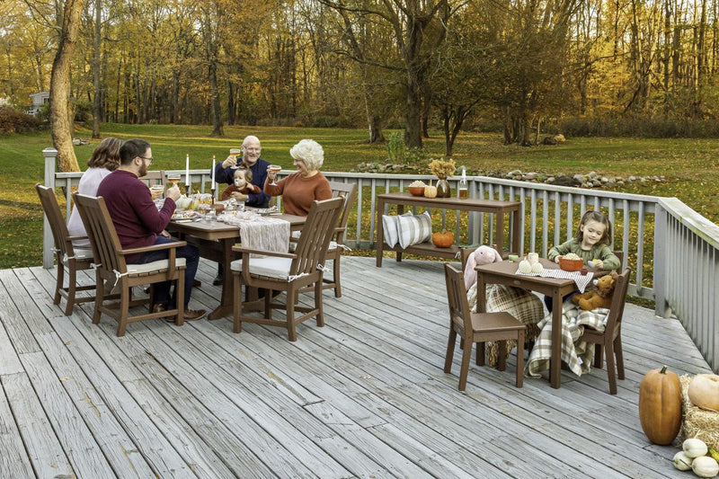A family gathers for a meal at assorted brown POLYWOOD dining sets on a wooden deck with fall decor by a lawn and tall trees.