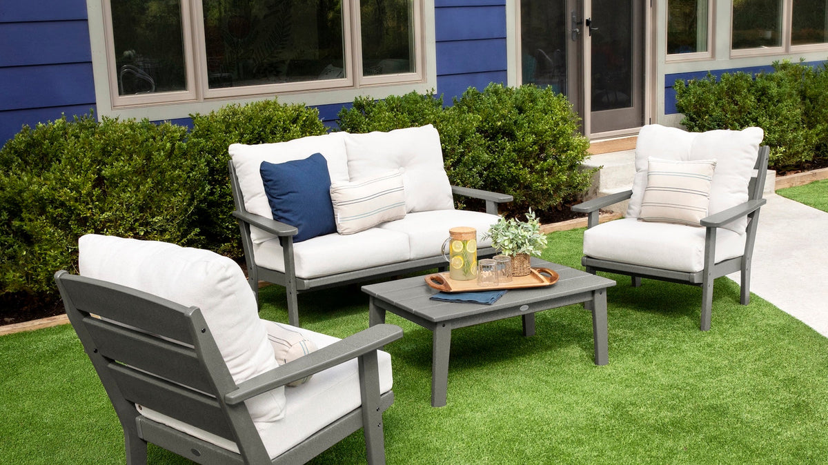 A POLYWOOD Lakeside 4 Piece Deep Seating Set in Slate Grey and Natural Linen is arranged on a green lawn before a blue house.