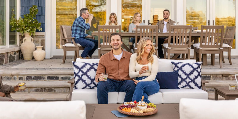 A man and a woman hold drinks and sit together on a tan PW Designer Series Estate Sofa with navy blue and white pillows.