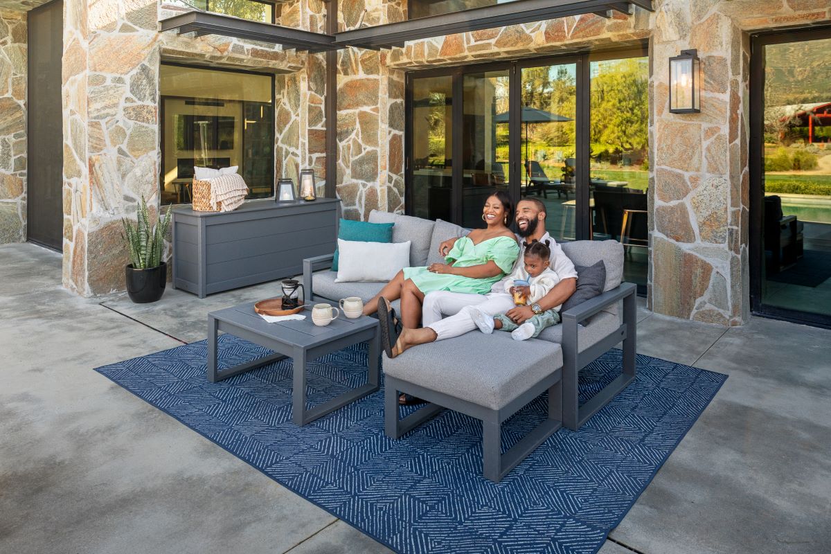A woman, man, and little girl relax together on a grey POLYWOOD EDGE sectional on a patio before a flagstone house.