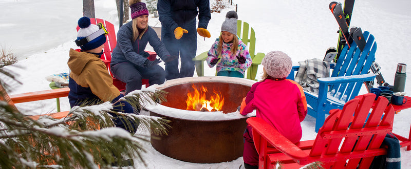 A snowy scene featuring a family bundled in coats and hats, seated on various POLYWOOD furniture around a fire pit.