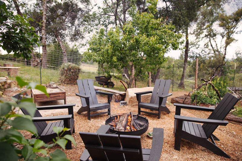 A group of black POLYWOOD Modern Adirondack Chairs sit around a fire pit in a rustic backyard surrounded by trees and fencing.