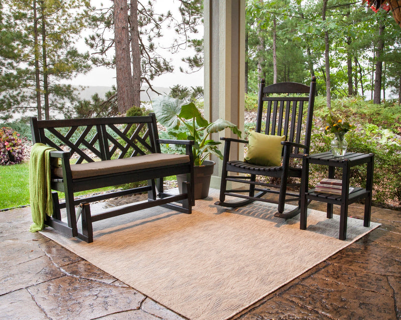 A black POLYWOOD glider bench, rocking chair, and side table sit on an area rug over a flagstone patio with tall trees in the background.