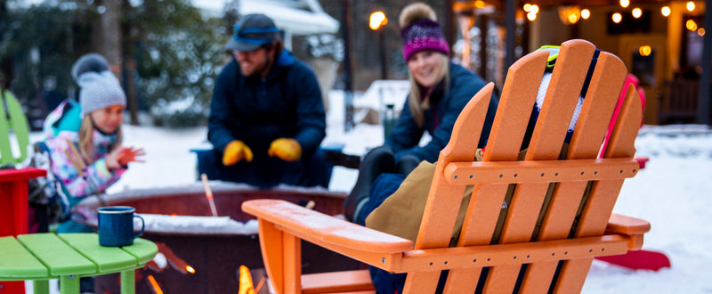 An orange POLYWOOD Classic Folding Adirondack Chair sits by a fire pit with people gathered around on a snowy day.