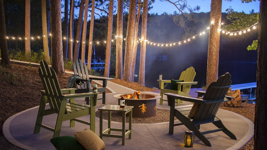 Assorted green POLYWOOD Long Island Adirondack chairs and side tables circle a fire pit in a wooded area by a lake.