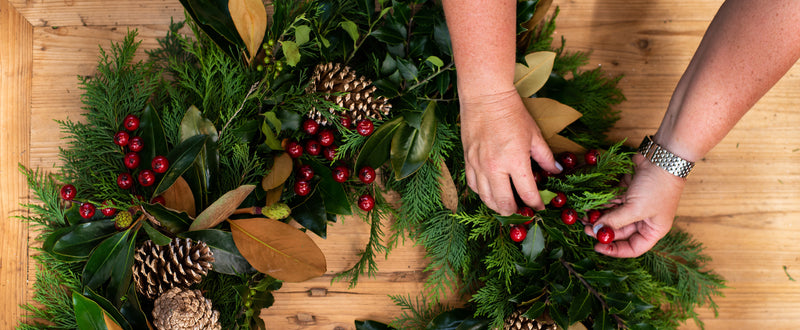 Hands adding red cranberries to a festive evergreen wreath decorated with pine cones and magnolia leaves.