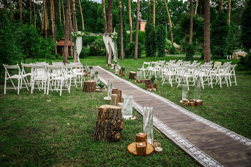 An outdoor wedding ceremony site is set up in a forest with white folding chairs, a long runner, an arbor, and woodsy decor.