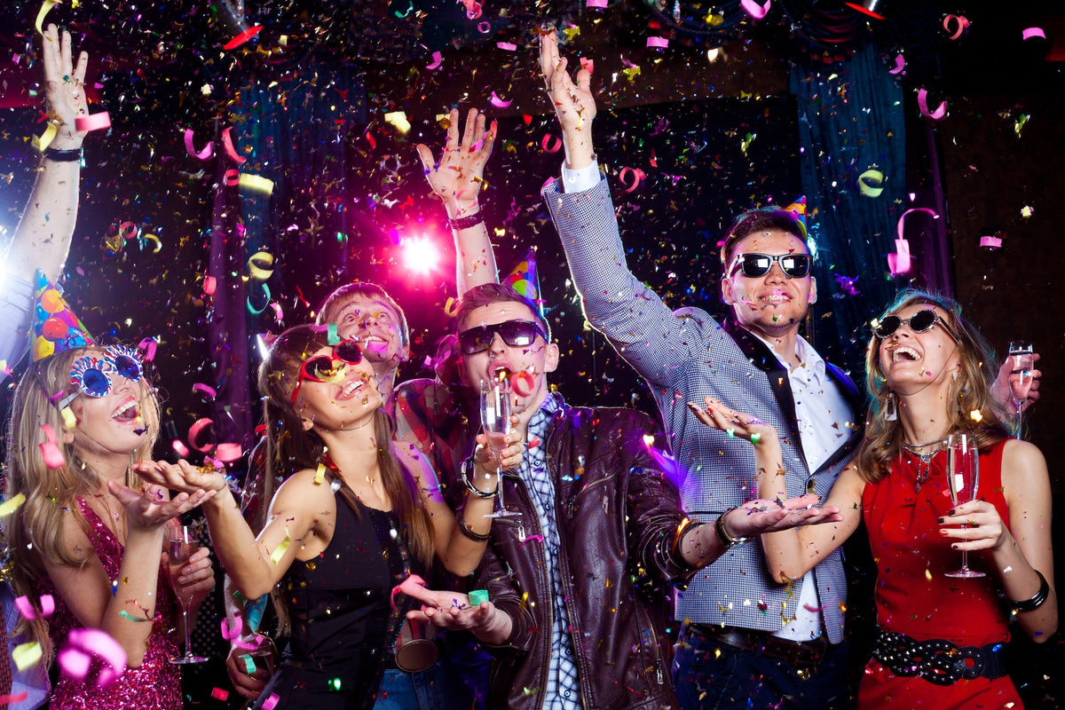 A group of people celebrating. They’re wearing sunglasses and party hats, and confetti is falling around them. 