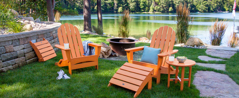 A Tangerine POLYWOOD Classic Oversized Adirondack 5-Piece Casual Set sits near a fire pit by a scenic lake enclosed by trees.