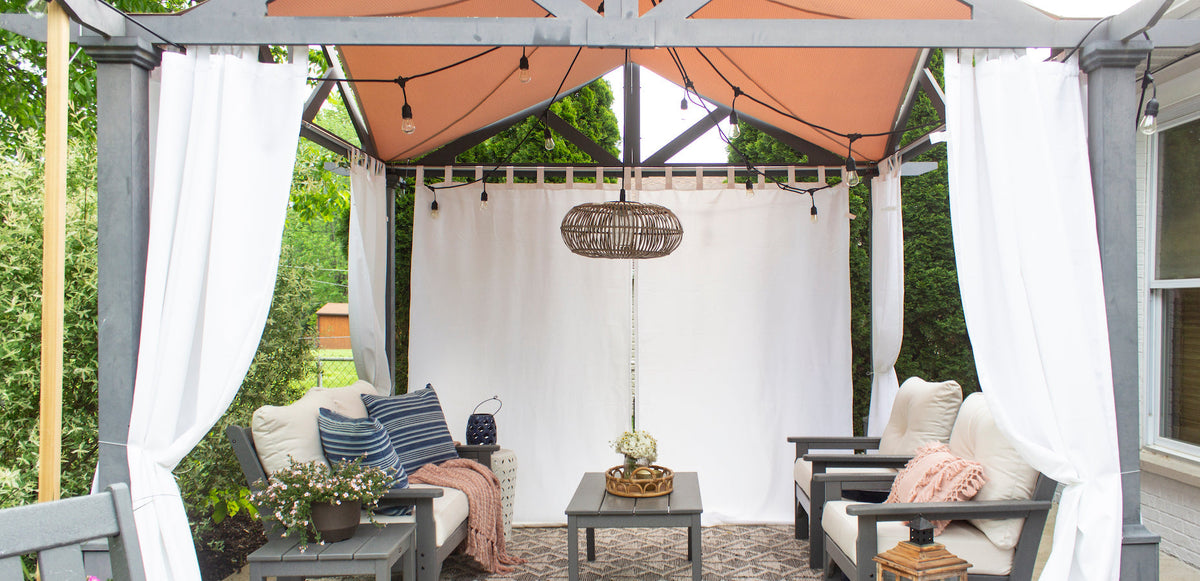 Assorted POLYWOOD deep seating furniture and tables sit beneath a grey pergola with white curtains and string lights.