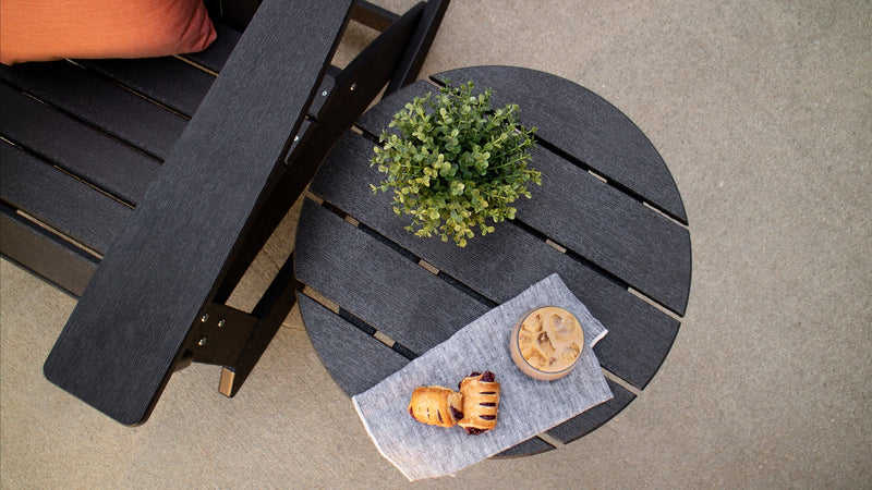 An overhead shot features a POLYWOOD Round 18 Inch Side Table in Black topped with a plant and a glass of iced coffee. 