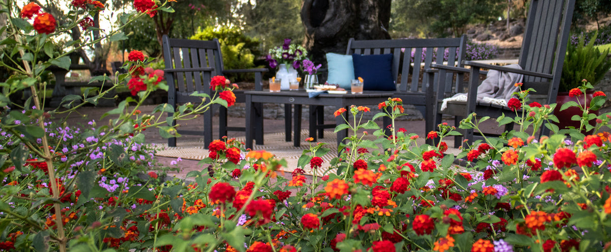 A POLYWOOD furniture set in Slate Grey sits on a patio with colorful flowers in the foreground.