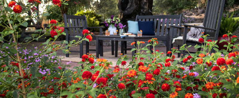 A POLYWOOD furniture set in Slate Grey sits on a patio with colorful flowers in the foreground.