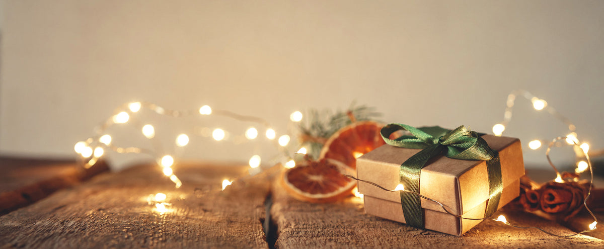 A small box tied with green ribbon sits on a wood surface with glowing fairy lights, dried fruit slices, and cinnamon sticks.