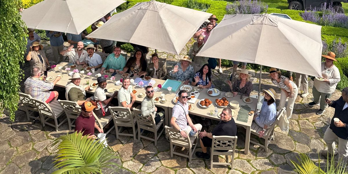 People gather at tan Martha Stewart patio sets by POLYWOOD, shaded by umbrellas on a stone patio surrounded by lush greenery.