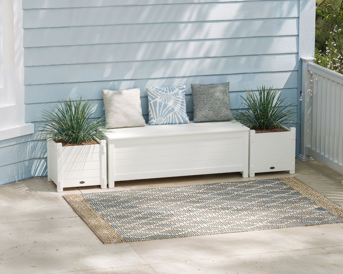 A white POLYWOOD 48 Inch Nautical Straight Planter Storage Bench Set sits beside an area rug on the balcony of a blue house.