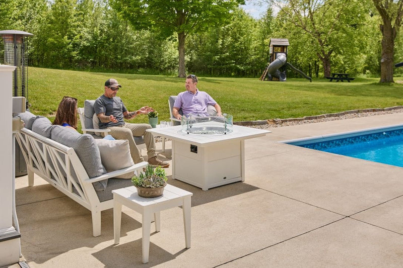 People chat on a poolside patio while relaxing in white POLYWOOD Braxton deep seating furniture by a fire pit table.