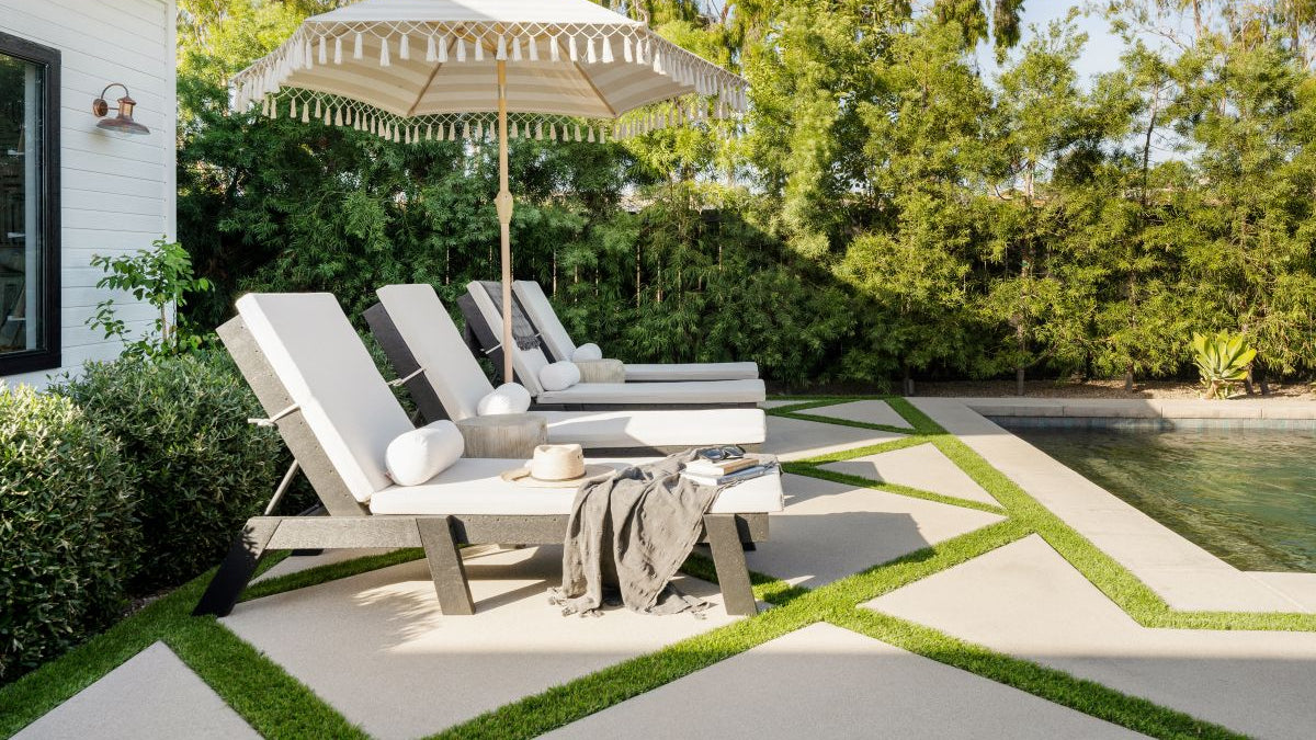 Black POLYWOOD Captain Chaises with cushions shaded by an umbrella line a paver patio by a pool with trees in the background.
