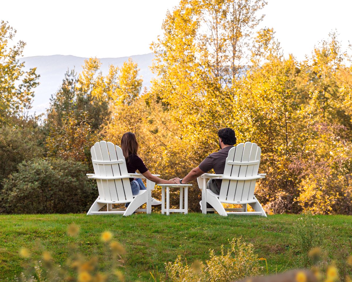 A couple holds hands while sitting at a POLYWOOD Vineyard 3 Piece Curveback Adirondack Set in White before a forest.