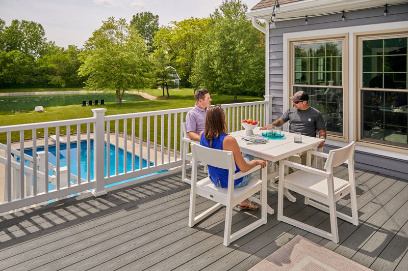 People sit at a white POLYWOOD EDGE 5 Piece Farmhouse Dining Set on a deck balcony overlooking a pool, lawn, and trees.